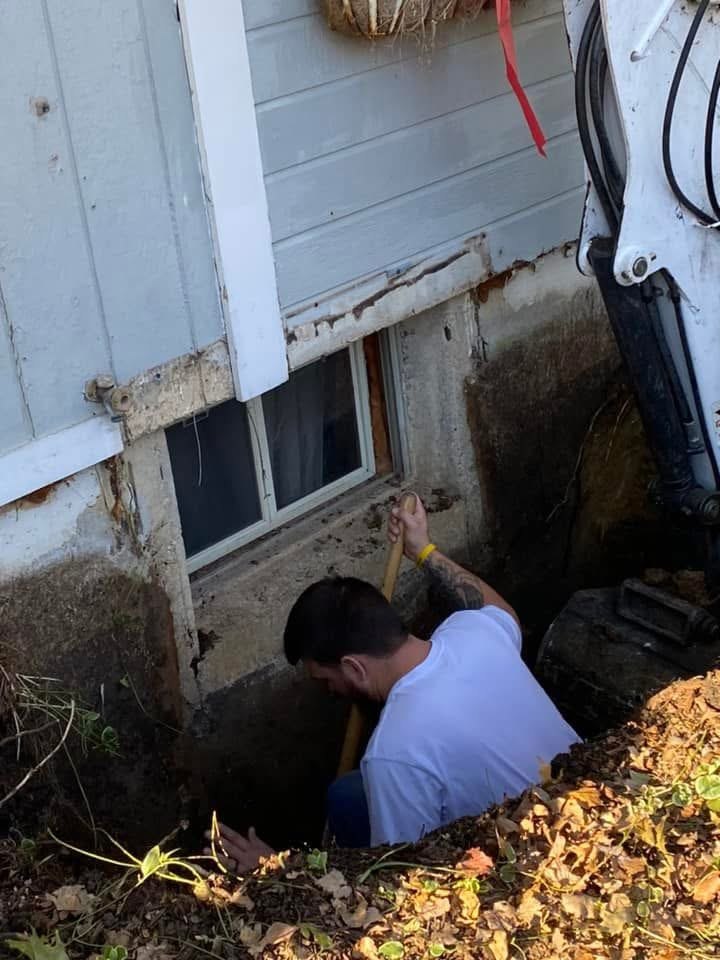 Hand-digging beside a basement window while an excavator stands by