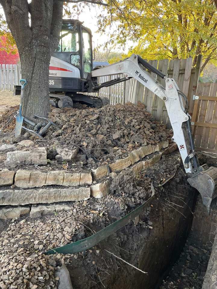Bobcat excavator cutting a deep trench beside a stone retaining wall in a Kansas backyard